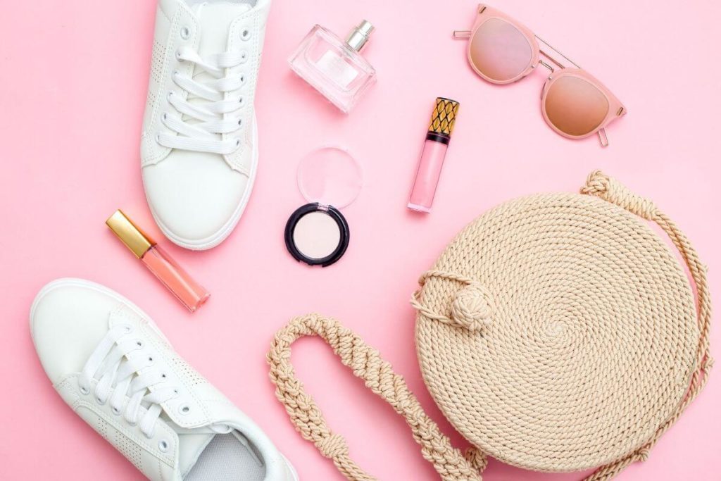 platte foto van een tas van stro, witte sneakers, roze lipgloss, roze oogschaduw, parfum en roze zonnebril.