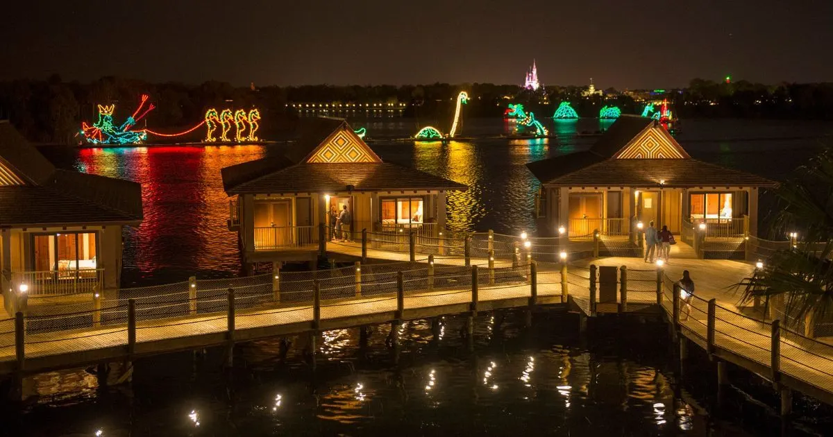 Horizontal image with Disney's Polynesian Village Resort bungalows in the foreground, the Electrical Water Pageant in the middle, and Cinderella's Castle in the background.