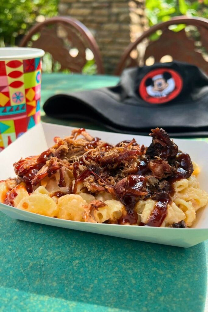 Photo of baked macaroni and cheese topped with barbecue pulled pork, with a Mickey Mouse club bucket hat and a cup of water from Disney World in the background.