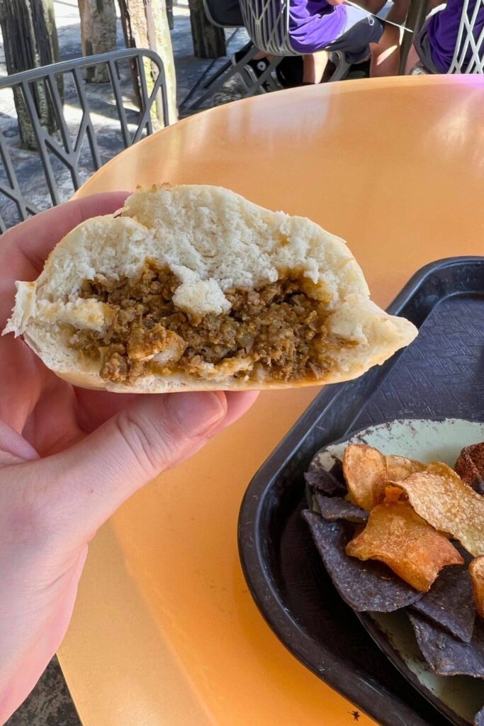 Photo of a steamed cheeseburger pod, aka a bao bun, cut in half, with a plate of chips in the background.