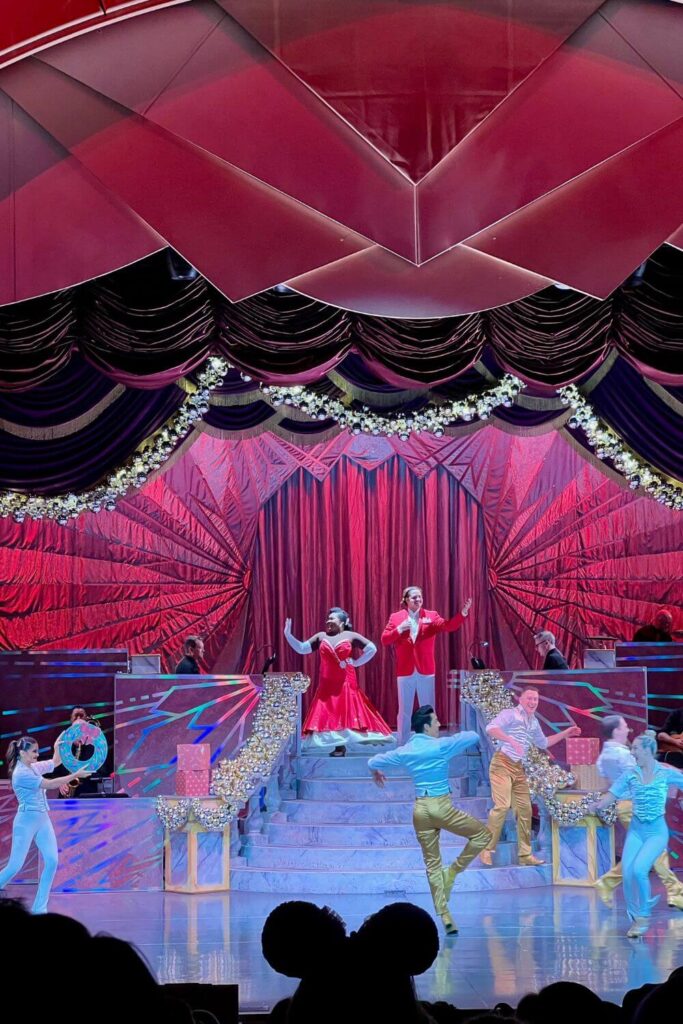 Photo of a man and woman at the top of stairs in fancy red holiday attire while men and women at the end of the stairs are dancing while wearing white and gold top and pants.