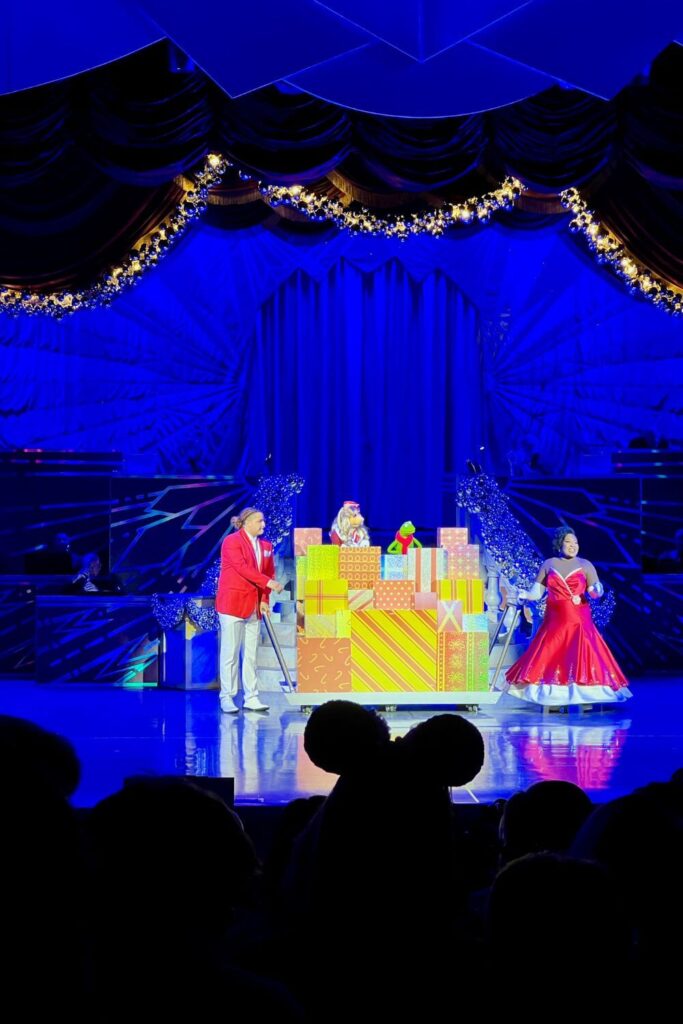 Photo of an opening scene of the Disney Holidays in Hollywood stage show at Jollywood Nights. On stage are the hosts, a male and female in red, fancy holiday attire, with a stack of Christmas gifts between them with Miss Piggy and Kermit the Frog puppets on top.